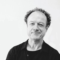 Black-and-white portrait of Niall Stokes with short, curly hair, wearing a black shirt over a black T-shirt, smiling slightly at the camera against a plain light background.