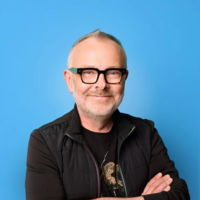 Portrait of Rick O’Shea wearing black glasses, a black T-shirt and a black quilted vest, smiling with his arms crossed against a bright blue background.