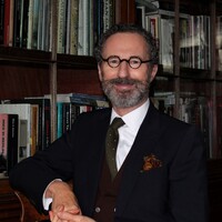 Portrait of Robert O'Byrne with curly salt-and-pepper hair, a full beard and round black glasses poses in front of wooden bookshelves filled with books. He wears a dark suit, white shirt, brown waistcoat, olive-green tie and patterned pocket square, smiling at the camera.