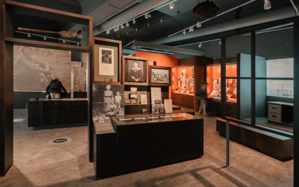 Section of 'Seamus Heaney: Listen Now Again' exhibition. A low brown case with black and white images of Seamus Heaney taken during his childhood and early adult years above it. He is shown graduating from college, with primary school classmates and with a family member. A visitor is shown stooped over looking at a different case in the background.