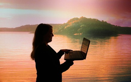 The profile of a woman is seen using a laptop against a picturesque background of a lake with land in the distance 