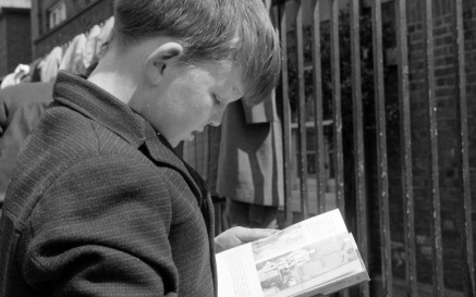 Léann buachaill óg i gcóta olla leabhar agus é ina sheasamh in aice le claí iarann saoirsithe 