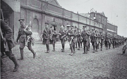 Soldiers in uniform with rifles on their shoulders march past a barracks in this black and white photograph