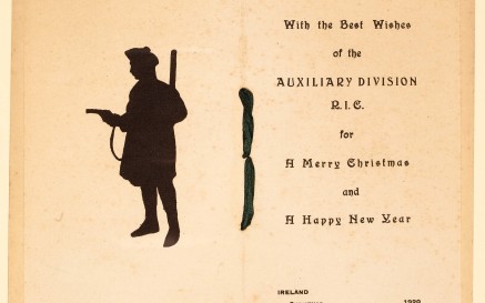 A yellowed greeting card bound by ribbon shows a silhouette of a soldier pointing a pistol, with text on the page opposite that reads: "With the best wishes of the Auxiliary Division R.I.C. for a Merry Christmas and a Happy New Year" "Ireland, Christmas, 1920"