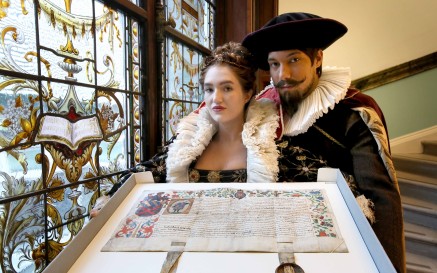 Two actors in period costume pose with grant of arms next to stained glass window