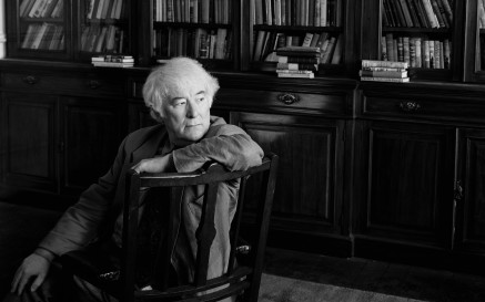 Grianghraf dubh agus bán de Sheamus Heaney ina shuí i gcathaoir agus seilfeanna leabhar ar a chúl