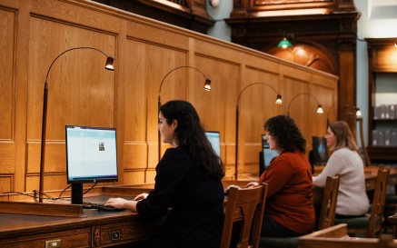 Suíonn triúr ban ag déanamh taighde ar scáileáin ríomhaire taobh istigh de phríomhsheomra léitheoireachta an NLI