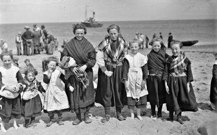 Grianghraf dubh agus bán de ghrúpa ban agus leanaí ar thrá ar Oileáin Árann ag caitheamh éadaí traidisiúnta