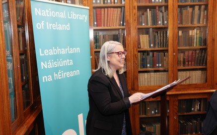 Woman speaking from a podium with bookshelves in the background