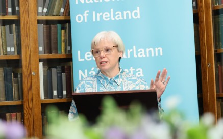 A woman giving a speech in front on a blue banner and bookshelves