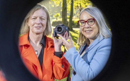 Two women holding a camera pointed at the viewer, with black circular frame 