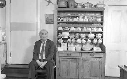 Seamus Heaney sitting in a wooden chair in a rural kitchen next to a cabinet filled with crockery