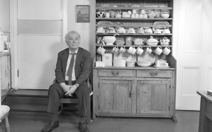Seamus Heaney sitting in a wooden chair in a rural kitchen next to a cabinet filled with crockery
