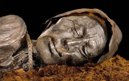 Close-up photograph of the preserved head of the Tollund Man, a naturally mummified body found in a Danish peat bog. His eyes are closed and his face appears peaceful, with detailed features and leathery skin preserved by the bog’s conditions. A rope noose is visible around his neck, and he wears a pointed leather cap.