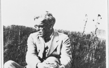 Black and white photograph of poet W. B. Yeats sitting in a grassy field. He is dressed in a light-colored suit and glasses, with his arms wrapped around one knee, gazing to his left. The setting is natural and serene, with tall grass and wild plants in the background.