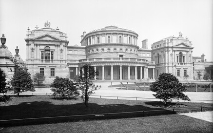 Black-and-white photograph of the National Library of Ireland in Dublin, showing its grand neoclassical facade. The central rotunda with tall columns and circular windows is flanked by two ornate rectangular wings topped with statues. In the foreground, landscaped gardens with trees and pathways frame the library building.