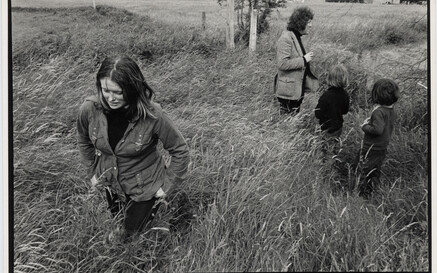 Black-and-white photograph of a young Marie Heaney walking through tall grass in a rural field, looking down as wind bends the grass around her. In the background, Seamus Heaney stands near a fence with two young children, all partially obscured by the grass, with open farmland stretching behind them.
