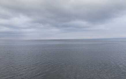 A wide view of the Flaggy Shore stretching to the horizon beneath a cloudy sky, with rounded grey stones in the foreground at the water’s edge.”