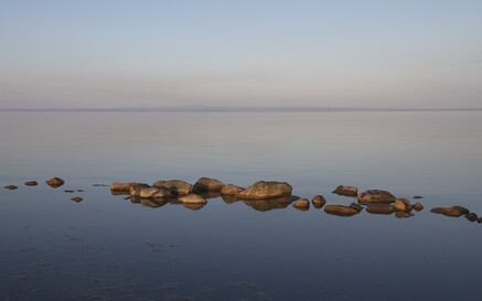Still water at Lough Neagh with a line of smooth rocks breaking the surface, reflected clearly in the calm lake. The horizon is faint in the distance beneath a wide, pale blue sky, creating a quiet, minimalist scene.