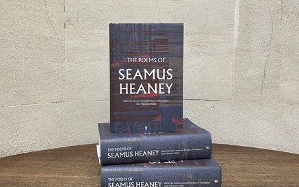 A stack of three copies of The Poems of Seamus Heaney sits on a wooden table against a pale stone wall. Two copies are laid flat with one standing upright on top, showing the book’s dark, textured cover design and title.