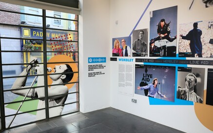 Music exhibition interior featuring large concert photos, including a singer on a window graphic and a wall display about Live Aid at Wembley, with text panels and a QR code.