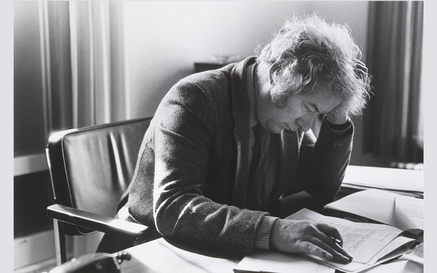 A black and white photo of Heaney writing poetry at his desk, holding his head in his hand