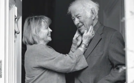 Black-and-white photo of Marie Heaney standing in a doorway, smiling warmly as she gently holds Seamus Heaney's face. Heaney, wearing a suit jacket, smiles back with one hand in his pocket, sharing an affectionate, intimate moment.