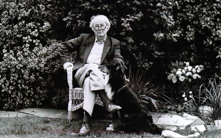 A black-and-white photo of Seamus Heaney sitting in a wicker chair in a garden. He wears a jacket, collared shirt, and trousers, sitting with one leg crossed over the other. Carlo the dog sits beside him, looking up at him. Dense leafy bushes fill the background, and the ground is grassy.