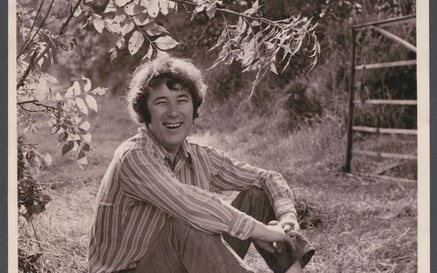 Black-and-white photograph of a young Seamus Heaney sitting on grass in a rural field, smiling at the camera. He wears a striped long-sleeve shirt, trousers, and lace-up boots, with his arms loosely wrapped around his knees. Tree branches with leaves hang above him, and a wooden gate stands in the background. 