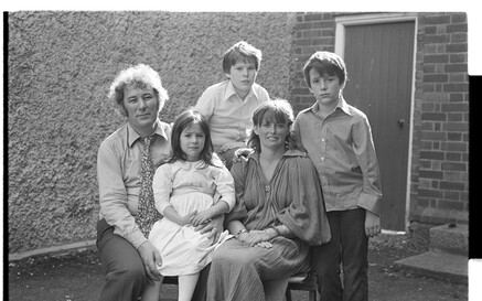 Black-and-white portrait of the Heaney family posing outdoors. Seamus and Marie sit on chairs in the center while their daughter Catherine Anne sits on Seamus’s lap. Their two sons, Michael and Christopher, stand behind and beside them. All face the camera with neutral expressions. The group is in a small paved yard with a rough-textured wall on the left and a brick wall with a door on the right.