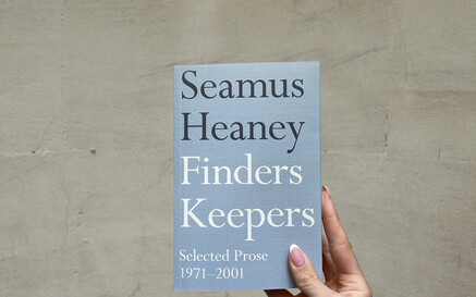 Hand holding a light blue copy of Finders Keepers by Seamus Heaney against a curved stone wall background.