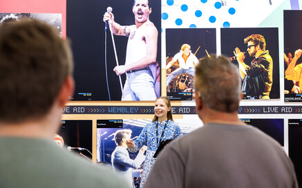 A female tour guide smiles and gestures while speaking to a small group of visitors in front of a large exhibition wall featuring iconic concert photographs. A prominent image of Freddie Mercury, shirtless and performing with a microphone dominates the display, with additional photos of other musicians arranged around it. The visitors are seen from behind, focused on the guide during the tour.